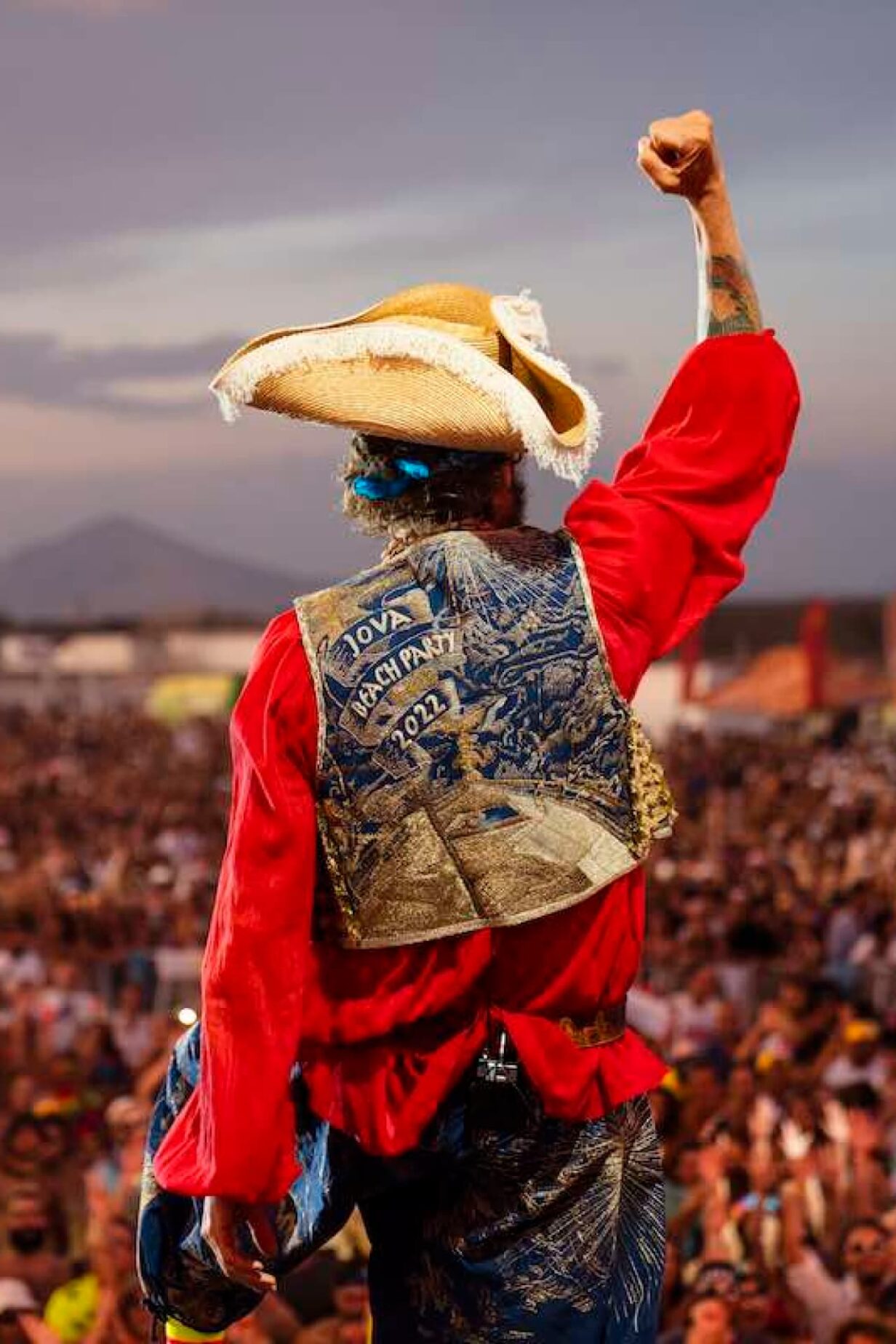 Jovanotti sul palco del Jova Beach Party 2022, visto di spalle, indossa un cappello, una camicia rossa e un gilet decorato. Davanti a lui, un'enorme folla.