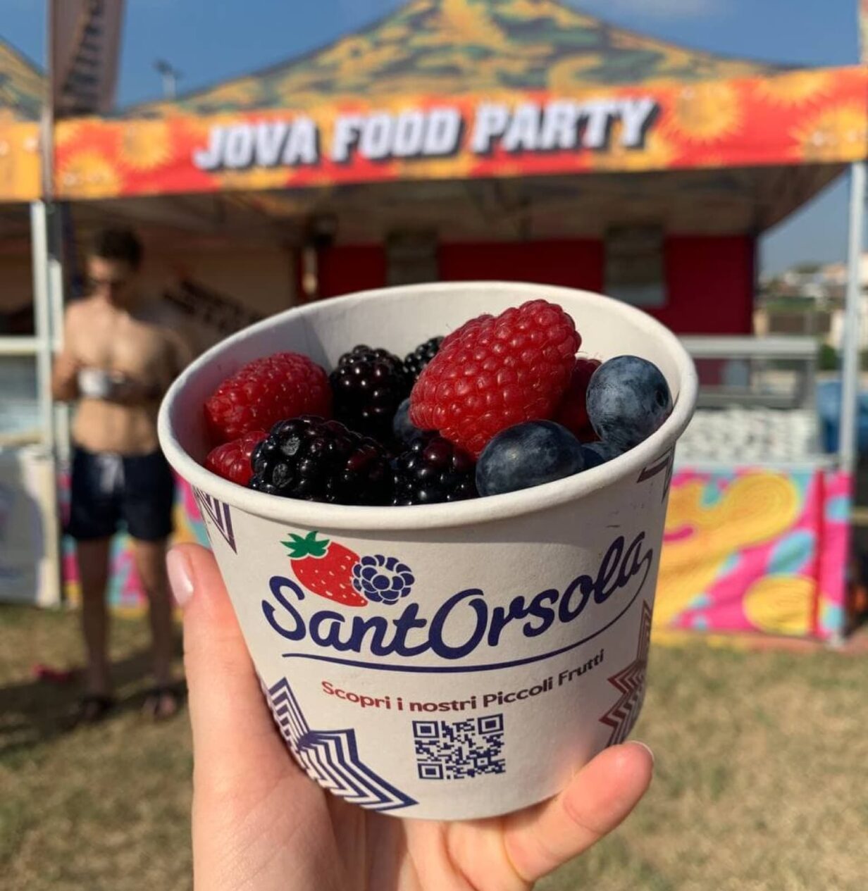 Due mani che tengono un contenitore di frutti di Sant'Orsola, con sullo sfondo uno stand del Jova Beach Party.