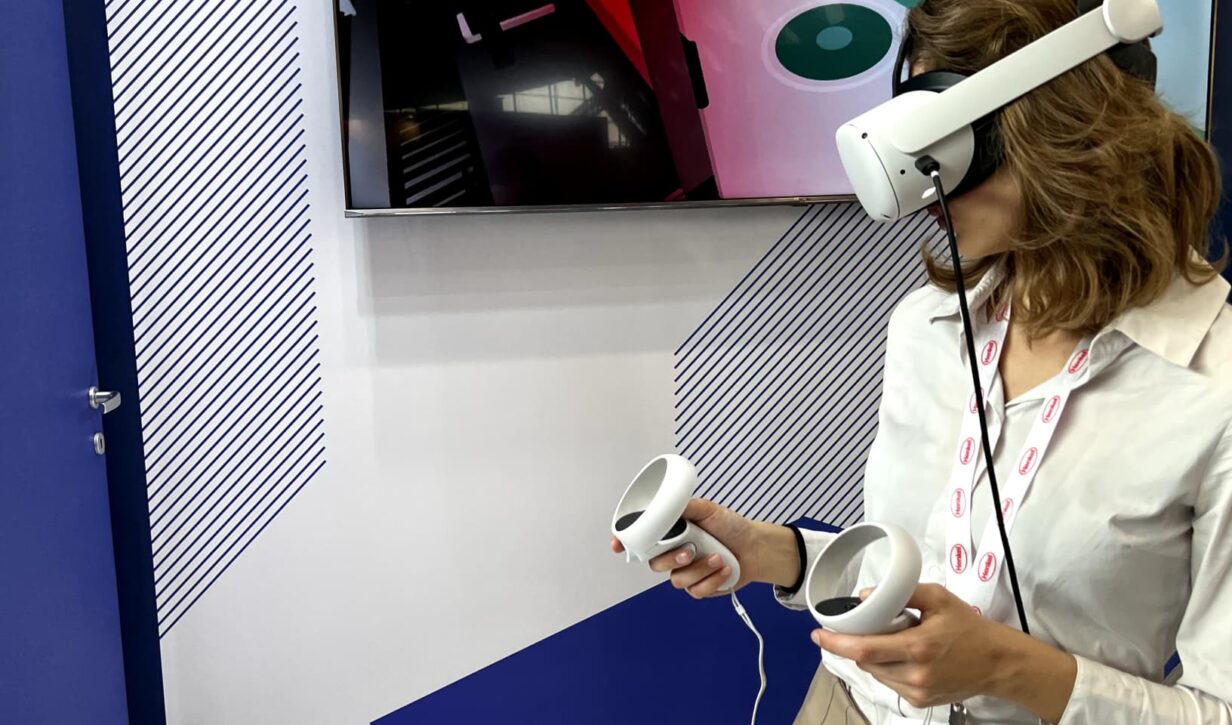 Donna con visore e controller nelle mani, coinvolta in un'esperienza virtuale immersiva durante un evento fieristico B2B.