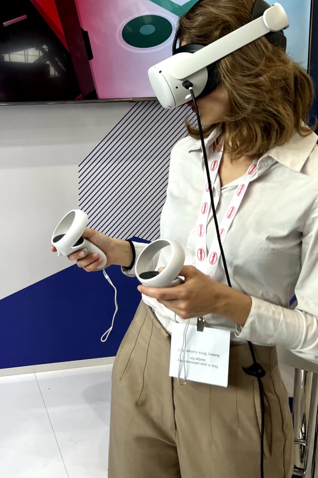 Donna con visore e controller nelle mani, coinvolta in un'esperienza virtuale immersiva durante un evento fieristico B2B.