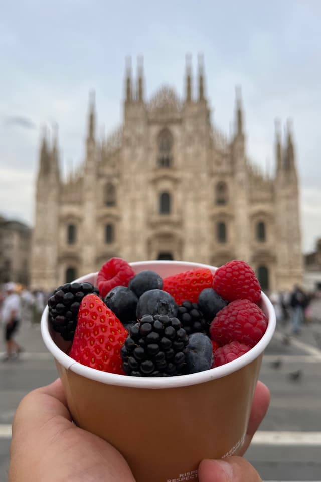 Una mano tiene un bicchiere pieno di frutti di bosco, tra cui fragole, lamponi, more e mirtilli. Sullo sfondo, il duomo di Milano sfocato.