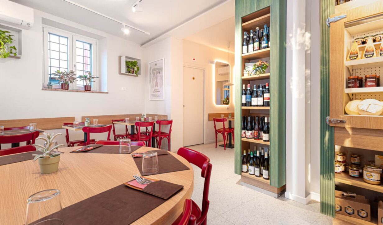 Sala interna del locale con tavoli in legno, sedie rosse e dettagli verdi. In evidenza una cantinetta con vini e prodotti gastronomici.
